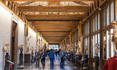 The Uffizi Gallery museum in Florence, Italy, with people walking through the hallways looking at sculptures and paintings on display. the ceiling is painted with frescoes. there's an art gallery-like atmosphere with bright natural light coming from large windows on both sides of the hallway