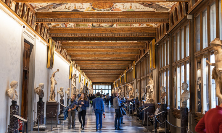 The Uffizi Gallery museum in Florence, Italy, with people walking through the hallways looking at sculptures and paintings on display. the ceiling is painted with frescoes. there's an art gallery-like atmosphere with bright natural light coming from large windows on both sides of the hallway